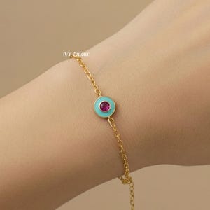 May include: Gold chain bracelet with a turquoise enamel circle and a ruby-coloured centre stone. The bracelet has an adjustable chain and a small charm. The brand name "IVY L'amour" is printed on the image.