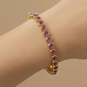 May include: A gold-colored bracelet with a series of round, magenta-colored gemstones. The bracelet is worn on a wrist, showcasing the jewelry's design and color. The gemstones are evenly spaced and set in a gold band.