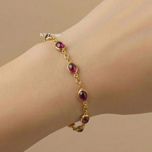 May include: A gold-colored bracelet featuring oval-shaped, deep red gemstones. The bracelet is composed of small links connecting the gemstones. The bracelet is worn on a wrist.