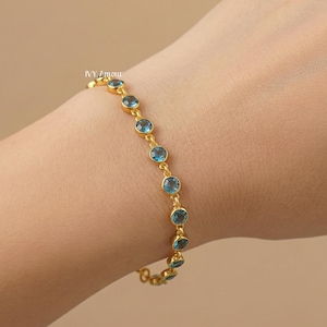May include: A gold-colored bracelet with round, light blue gemstones. The bracelet is delicate and features a series of evenly spaced stones. The bracelet is on a wrist, against a neutral background.