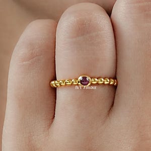 May include: A gold-colored ring with a beaded band and a small, round, dark red gemstone. The ring is on a finger, and the background is blurred. The ring is a delicate piece of jewelry.