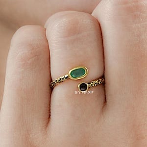 May include: A gold-toned ring with an open design, featuring an oval green stone and a round black stone. The ring has a textured band and is worn on a finger. The text "IVY Amour" is visible on the ring.