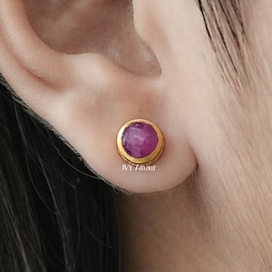 May include: A close-up of a gold stud earring with a round, pink gemstone. The earring is set in a gold bezel and is worn on an ear. The image is well-lit, showcasing the earring's design and color.