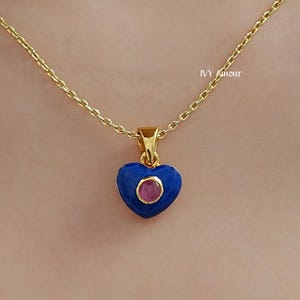 May include: A gold-colored necklace with a heart-shaped pendant. The pendant is blue with a small, round, red center. The necklace is delicate and appears to be worn around the neck.