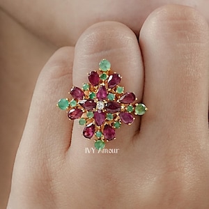 May include: A gold-toned ring featuring a floral design with ruby and emerald gemstones. The ring's center has a small diamond. The ring is worn on a finger. The ring is a piece of jewelry.