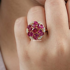 May include: A gold ring featuring a cluster of oval-cut, deep red gemstones surrounded by smaller, clear stones. The ring's design is a dome shape, with the gemstones set closely together. The ring is on a finger, with the brand name "NY 7mour" visible.