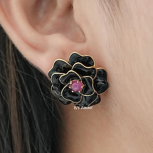 May include: Black flower-shaped stud earrings with a gold trim and a pink center stone. The earrings are worn on an ear. The earrings have a glossy finish and are likely made of metal.