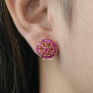May include: Close-up of a gold-toned stud earring with a cluster of small, round, ruby-colored stones. The stones are arranged in a circular pattern, creating a floral design. The earring is set against the backdrop of a person's ear and skin.