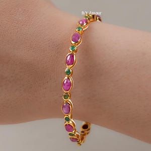 Puede incluir: Pulsera dorada con piedras preciosas rosas ovaladas y verdes redondas alternadas. La pulsera se lleva en la muñeca, mostrando el diseño y el contraste de colores de la joya. La pulsera es una pieza de joyería de moda.