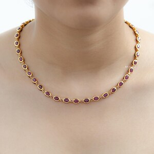 Op de afbeelding: Een goudkleurige ketting met ovale, dieprode edelstenen. De edelstenen zijn gezet in individuele zettingen en met elkaar verbonden tot een delicate ketting. De ketting wordt om de nek van een persoon gedragen.
