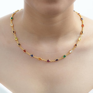 Pode incluir: Um colar dourado com uma série de pedras preciosas ovais e coloridas. As pedras são engastadas numa corrente delicada, criando uma joia vibrante. O colar é usado ao redor do pescoço.