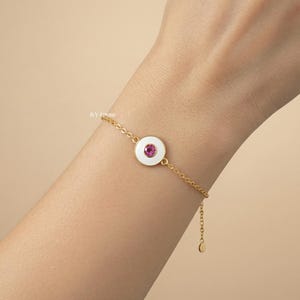 Puede incluir: Una pulsera dorada con un colgante circular blanco con una gema rosa en el centro. La pulsera tiene un dise&ntilde;o de cadena delicada y un cierre ajustable. La joya se muestra sobre un tono de piel claro.