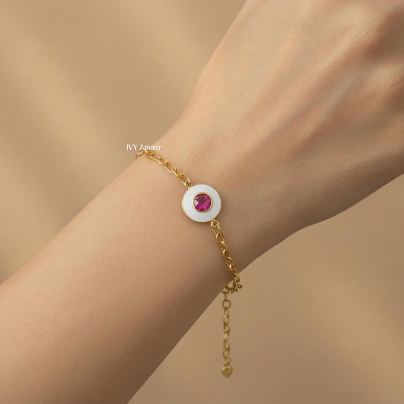 Puede incluir: Pulsera dorada con un dije circular blanco y una piedra central rosa brillante. La pulsera tiene un dise&ntilde;o de cadena delicado y se muestra en una mu&ntilde;eca.