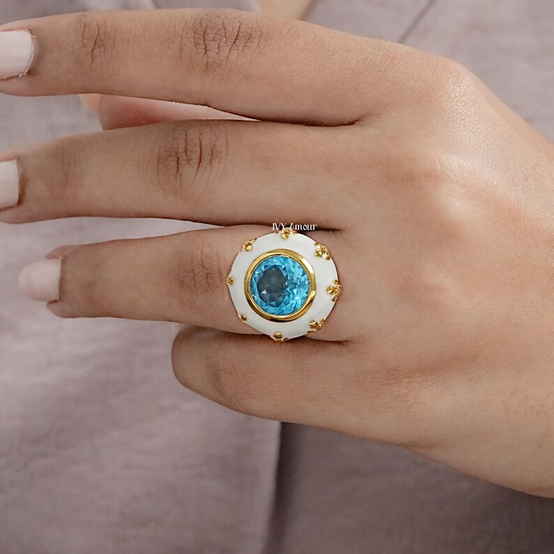 May include: A gold ring with a large, round, light blue gemstone set in a white enamel setting. The ring has gold accents around the white enamel. The ring is on a person's finger.