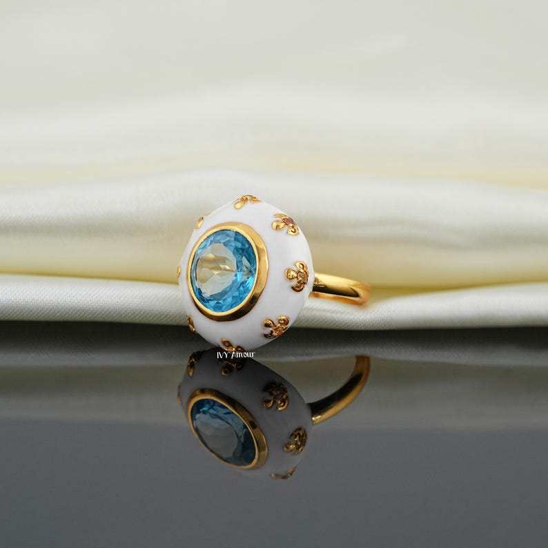 May include: A gold ring featuring a white, egg-shaped centerpiece with a blue gemstone. The ring has gold accents and floral designs. The ring is displayed on a reflective surface.