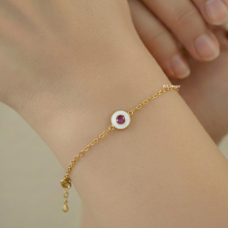 Puede incluir: Pulsera de cadena dorada con un c&iacute;rculo de esmalte blanco y una peque&ntilde;a gema roja redonda en el centro. La pulsera est&aacute; en una mu&ntilde;eca. La cadena tiene un dise&ntilde;o delicado.
