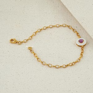 Puede incluir: Pulsera de cadena dorada con un dije circular blanco y rosa. El dije tiene una piedra central rosa y un borde blanco. La pulsera tiene un cierre de mosquet&oacute;n.