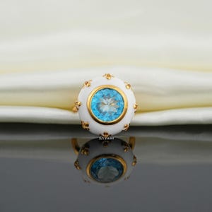 May include: A gold ring featuring a large, round, light blue gemstone set in a white enamel setting. The setting is accented with gold details. The ring is reflected in a dark, glossy surface.