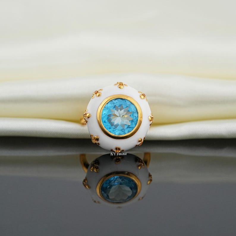 May include: A gold ring featuring a large, round, light blue gemstone set in a white enamel setting. The setting is accented with gold details. The ring is reflected in a dark, glossy surface.