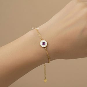 Puede incluir: Una pulsera dorada con un dije de esmalte blanco y una gema p&uacute;rpura en el centro. La pulsera es delicada y presenta un dise&ntilde;o de cadena. El dije es circular y la gema es de color p&uacute;rpura oscuro.