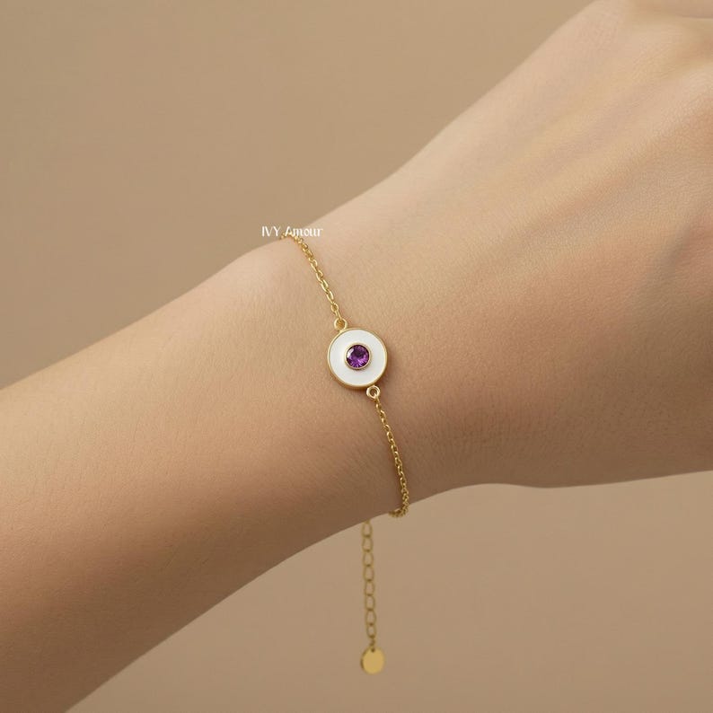 Puede incluir: Una pulsera dorada con un dije de esmalte blanco y una gema p&uacute;rpura en el centro. La pulsera es delicada y presenta un dise&ntilde;o de cadena. El dije es circular y la gema es de color p&uacute;rpura oscuro.