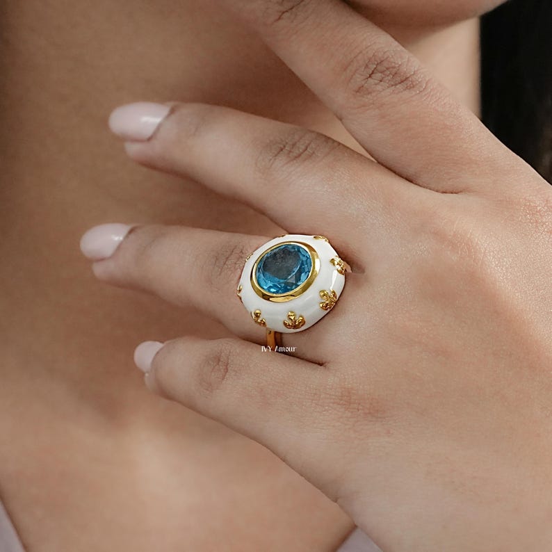 May include: A gold ring featuring a large, round, light blue gemstone set in a white enamel setting. The ring has gold accents and a delicate design. The ring is on a person's finger.