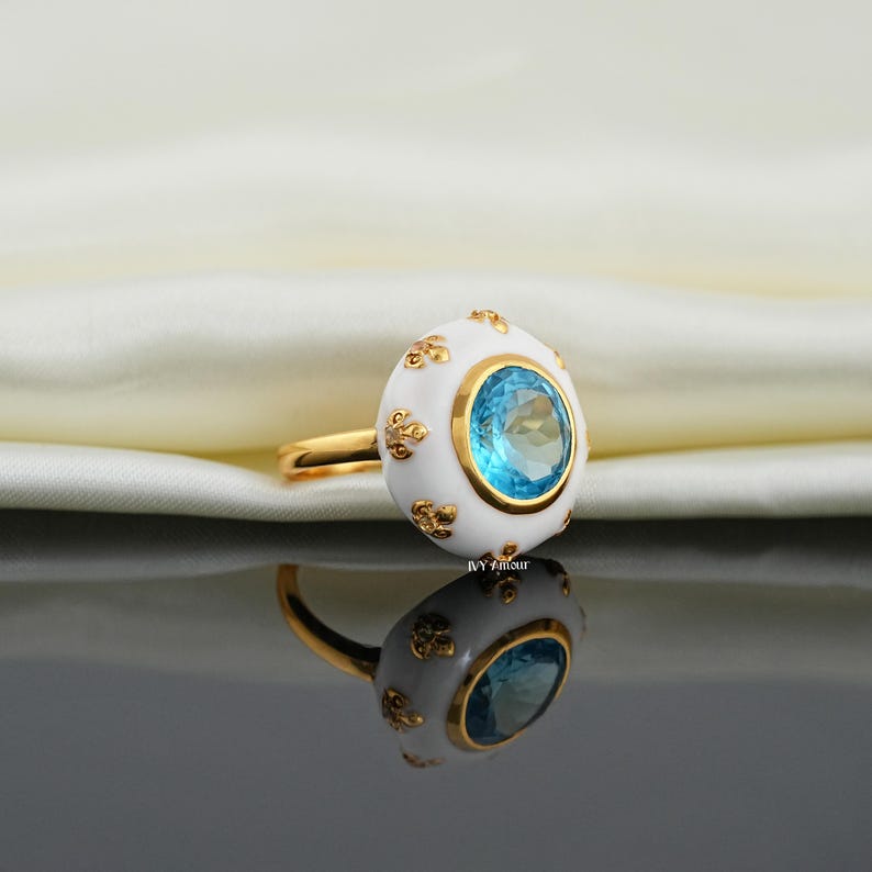 May include: A gold ring with a white enamel setting and a large, oval, light blue gemstone. The ring's setting is decorated with small gold floral accents. The ring is displayed on a reflective surface.