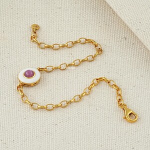 Puede incluir: Pulsera de cadena dorada con un dije circular blanco y rosa. El dije presenta una gema rosa en el centro y un borde de esmalte blanco. La pulsera tiene un cierre de mosquet&oacute;n y una peque&ntilde;a etiqueta decorativa.