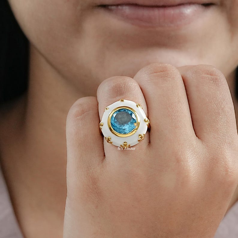 May include: A gold ring with a large, round, light blue gemstone set in a white and gold bezel. The ring has a unique, ornate design with small gold accents around the white border. The ring is on a person's finger.