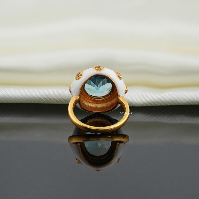 May include: A gold ring with a large, light blue gemstone. The ring features a white enamel setting with small gold accents. The ring is reflected on a dark, glossy surface.