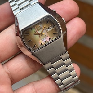May include: A vintage Orient automatic wristwatch with a silver-tone case and band. The watch face has a mother-of-pearl effect with gold hands and markers. The day is displayed as "FRI 31".