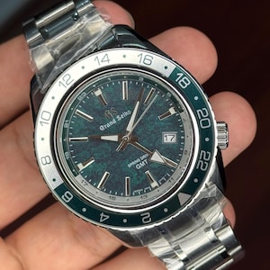 May include: A stainless steel Grand Seiko watch with a green dial and a green and silver bezel. The watch features a date window and the text "SPRING DRIVE GMT". The watch is on a stainless steel band.