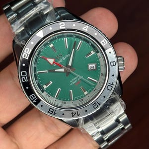 May include: A stainless steel wristwatch with a green dial and a silver bezel. The watch face displays the brand name "Grand Seiko" and the text "AUTOMATIC HI-BEAT 36000". The watch has a date display at the 3 o'clock position.