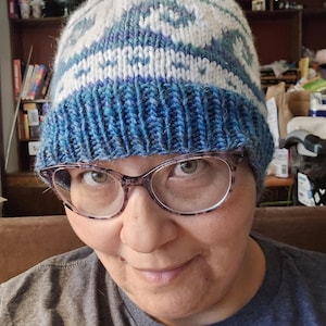 May include: A hand-knitted beanie featuring a ribbed blue brim and a crown decorated with a white and blue geometric pattern. The hat is constructed from yarn in shades of deep blue, light blue, and white, showcasing a textured knit design.