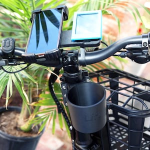 May include: Black bicycle handlebars with a phone mount, a small blue device, and a black cup holder. A black basket is attached below the handlebars. The background includes green palm leaves and a potted plant.