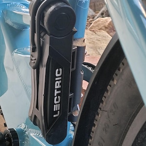 May include: A black Lectric bike lock is mounted on a light blue bicycle frame. The lock is secured to the frame with a black bracket. The bicycle tire is black with a 20 x 3.0 size.