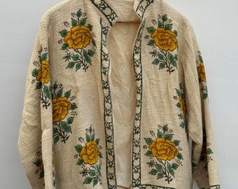 May include: Beige open-front jacket with yellow rose floral embroidery and green trim. The jacket is hanging on a wooden hanger. The jacket has a collarless design and long sleeves. The floral design is repeated across the jacket.