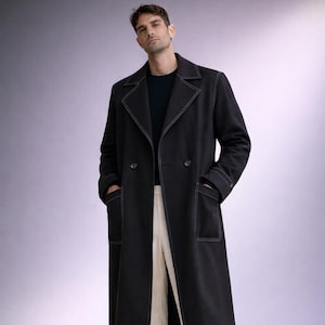 May include: A long, black coat with white stitching details and a double-breasted design. The coat is worn with cream-colored pants and black shoes. The coat has pockets and a classic, tailored look.