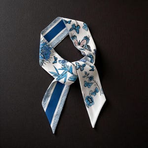 May include: A white silk scarf featuring a blue floral and butterfly design. The scarf has blue stripes and a blue floral pattern. The scarf is knotted, with the ends pointing downwards. The background is black.