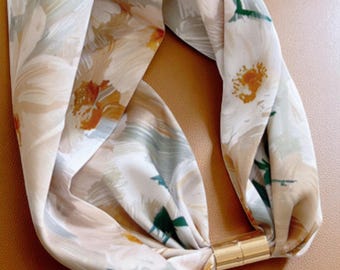 May include: A floral patterned headband with a gold-colored metal clasp. The headband features a cream-colored background with watercolor-style flowers in shades of orange, green, and brown. The fabric is twisted at the front.