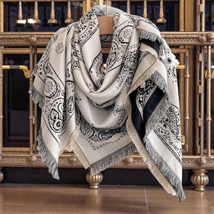 May include: Cream-colored scarf with a black paisley pattern and fringe trim. The scarf is draped over a wooden hanger, showcasing its intricate design. The scarf's edges are finished with a delicate fringe.