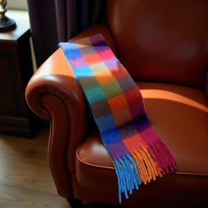 Puede incluir: Una bufanda a cuadros multicolor con borde de flecos, sobre un sillón de cuero marrón. La bufanda presenta un patrón vibrante de cuadrados en tonos de azul, verde, naranja, morado y rosa. La franja es una mezcla de azul y naranja.