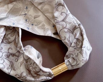 May include: A cream-colored headband with a gold-colored metal clasp. The fabric is patterned with a floral and bird design in shades of brown and gray. The headband is folded and looped, showcasing the pattern.