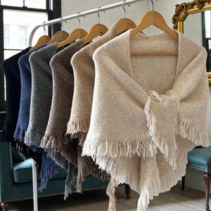 May include: A selection of shawls in shades of black, navy, grey, brown, and beige, displayed on wooden hangers. Each shawl features a fringed trim. The shawls are presented on a metal clothes rail.