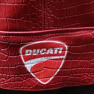 Op de afbeelding: Een felrode bucket hat met een gestructureerd krokodillenleerpatroon over het gehele materiaal. In het midden aan de voorzijde bevindt zich een wit schildvormig embleem met het woord DUCATI in dikke zwarte hoofdletters gedrukt.