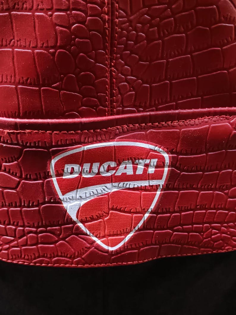 Op de afbeelding: Een felrode bucket hat met een gestructureerd krokodillenleerpatroon over het gehele materiaal. In het midden aan de voorzijde bevindt zich een wit schildvormig embleem met het woord DUCATI in dikke zwarte hoofdletters gedrukt.