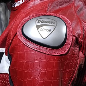 Op de afbeelding: Een rode motorjas met een gestructureerd krokodillenleerpatroon. De jas is voorzien van een zilverkleurige schouderbeschermer met de zwarte tekst DUCATI CORSE erop gedrukt. De kraag toont de witte tekst DUCATI op een rode achtergrond.