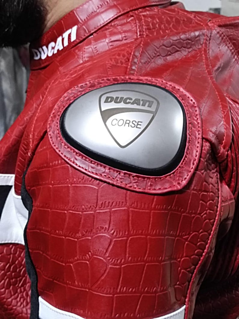 Op de afbeelding: Een rode motorjas met een gestructureerd krokodillenleerpatroon. De jas is voorzien van een zilverkleurige schouderbeschermer met de zwarte tekst DUCATI CORSE erop gedrukt. De kraag toont de witte tekst DUCATI op een rode achtergrond.