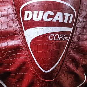 Op de afbeelding: Een rode jas met een gestructureerd krokodillenleerpatroon en een groot wit schildvormig logo op de achterkant. Het logo bevat de tekst DUCATI in vetgedrukte letters boven het woord CORSE, met witte biezen zichtbaar langs de onderste randen.