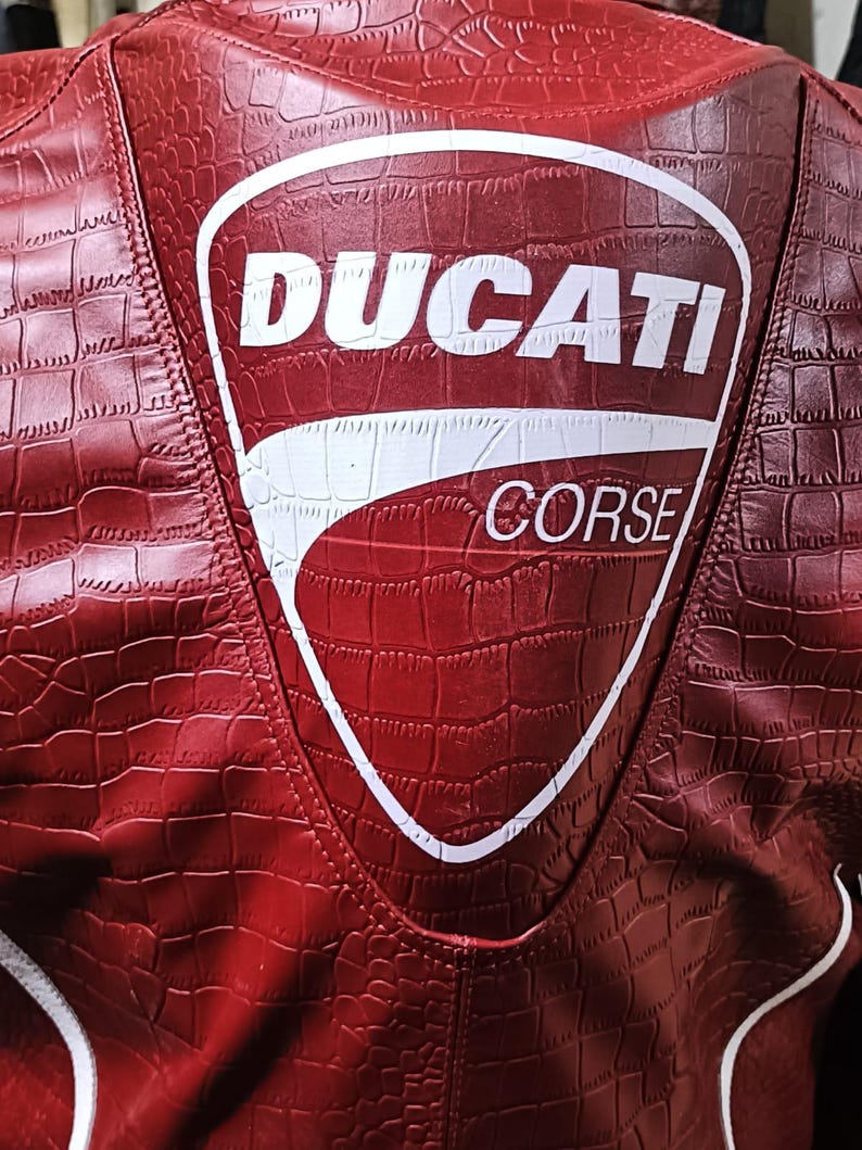 Op de afbeelding: Een rode jas met een gestructureerd krokodillenleerpatroon en een groot wit schildvormig logo op de achterkant. Het logo bevat de tekst DUCATI in vetgedrukte letters boven het woord CORSE, met witte biezen zichtbaar langs de onderste randen.